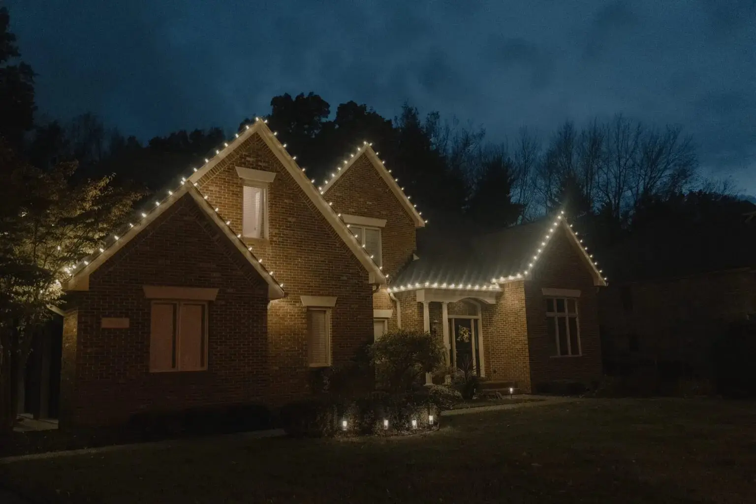 A brick house outlined in warm white holiday lights at dusk